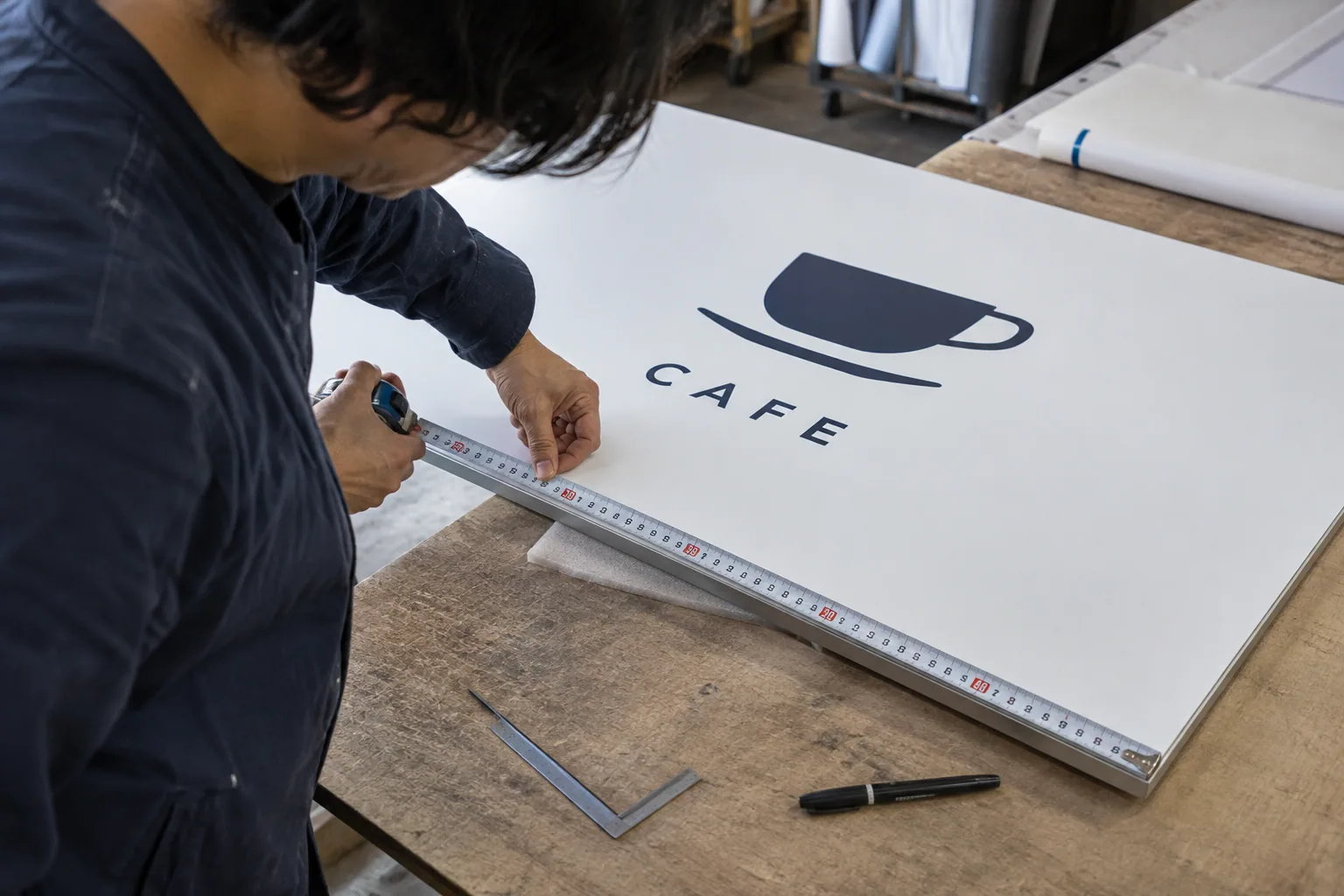 作業台でカフェの看板パネルを測る職人の生成イメージ